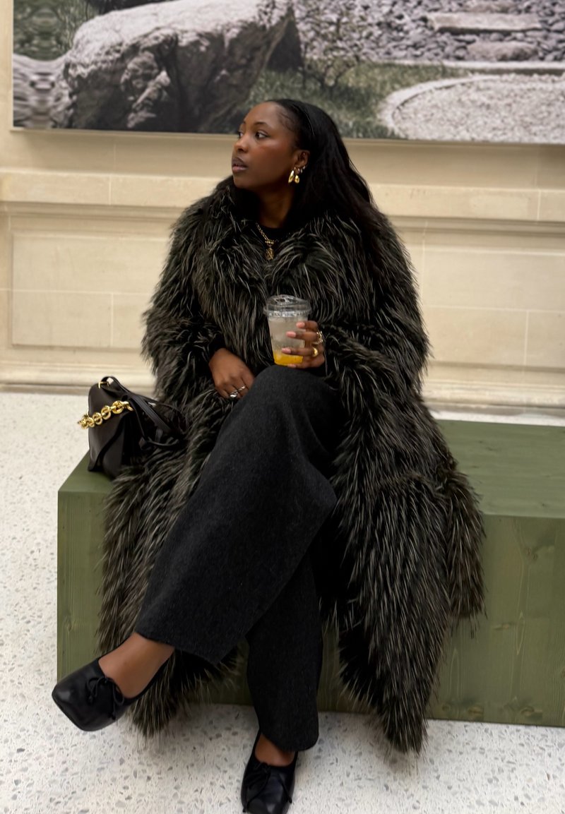 Femme en long manteau de fourrure noire assise sur un banc, tenant un verre, avec un sac à main noir à ses côtés et une grande photo en noir et blanc derrière elle.