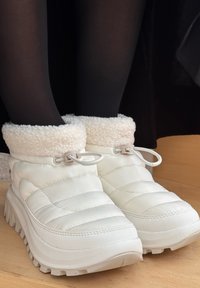 Botas de tobillo blancas, acolchadas, con un suave collar de crema, cordones elásticos y una suela gruesa, que presentan una superficie texturizada y una forma redondeada.
