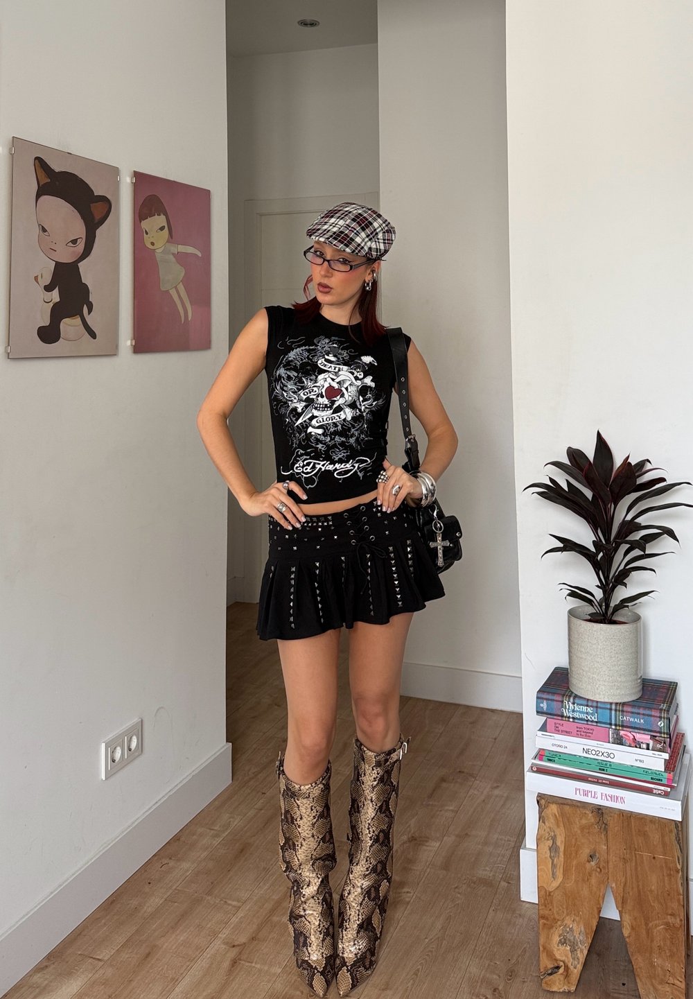 Femme portant un haut sans manches noir à motifs graphiques, une mini-jupe noire ornée de clous, des bottes mi-mollets en peau de serpent et une casquette à carreaux, se tenant à l'intérieur près d'une plante et d'une œuvre d'art.