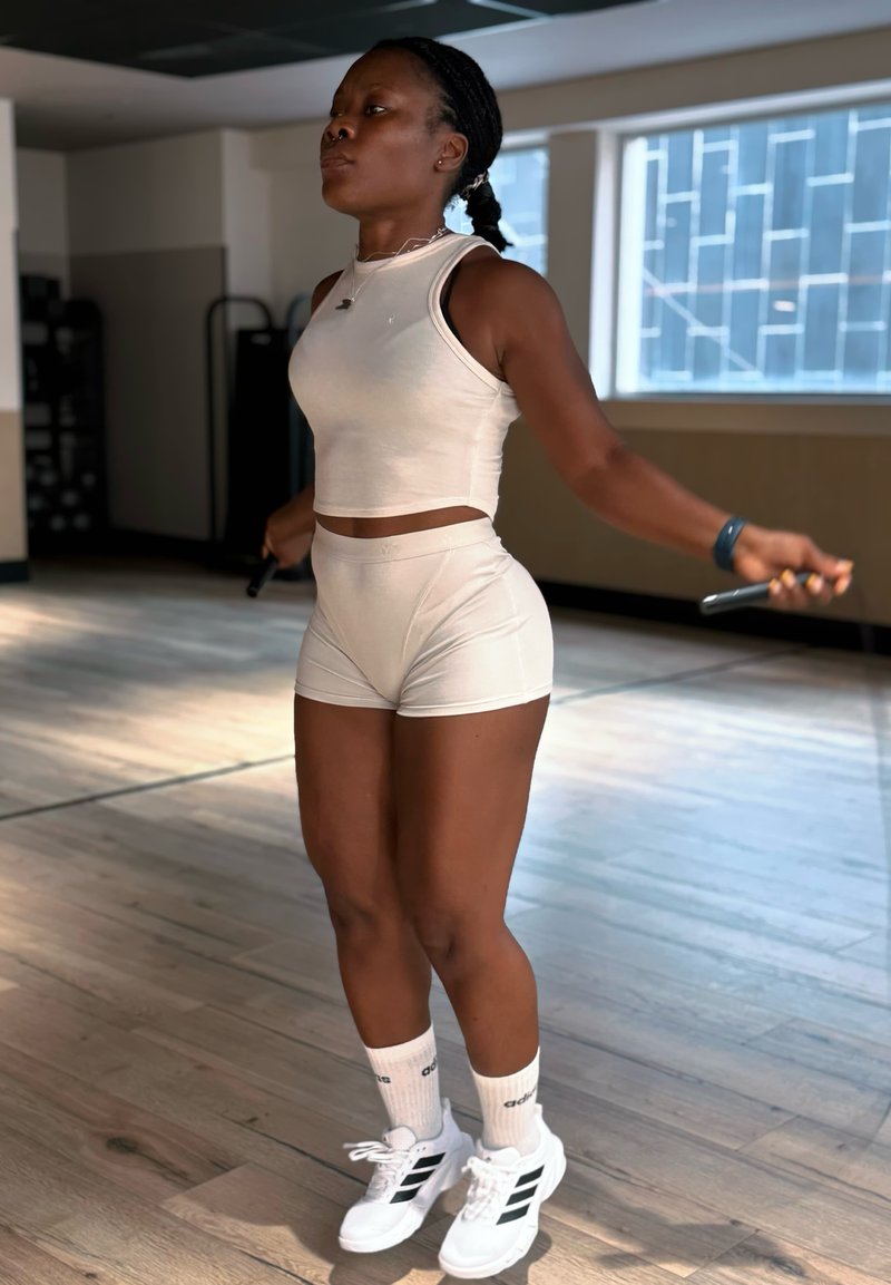Femme en tenue de sport blanche sautant à la corde dans une salle de sport avec des sols en bois et de grandes fenêtres.
