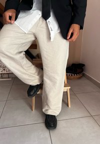 Μπεζ παντελόνια με φαρδιά πόδια από υφασμάτινο υλικό, συνδυασμένα με μαύρο σακάκι και μαύρα παπούτσια. Το πουκάμισο από κάτω είναι λευκό με κλειστό γιακά.