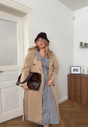 Mujer con gabardina beige, sombrero tipo bucket negro y vestido a cuadros sosteniendo un gran bolso marrón en una habitación minimalista con suelo de madera.