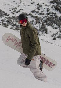 Snowboard med blomstrete rosa design holdt av en person i grønn jakke og hvite bukser. Svart hjelm og store reflekterende goggles brukt.