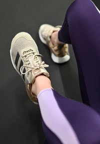 Chaussures de sport beiges avec dessus en mesh et rayures foncées, associées à des leggings violets et lilas, sur fond noir.