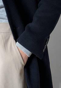 Personne portant un pantalon beige et un manteau noir avec quatre boutons sur la manche, la main reposant dans la poche, le poignet de la chemise gris clair visible.