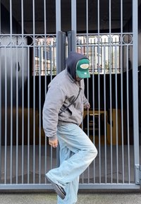 Giacca in pile grigia con zip, jeans azzurri e calzature nere. Persona passa attraverso un cancello metallico con barre verticali, parzialmente aperto.