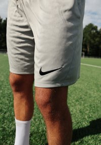 Nike sportnadrág világosszürke anyagból, fekete logóval a combon, fehér zoknikkal viselve egy zöld fűtéri pályán.