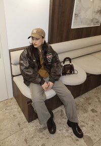 Νεαρός που φοράει καφέ καπέλο με σχέδια, σκούρο μπουφάν Adidas, μπεζ παντελόνι και μαύρες μπότες, κάθεται σε έναν πάγκο με κρεμ μαξιλάρι και κρατάει καφέ τσάντα.