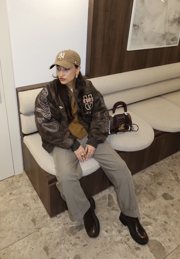 Noor inimene, kes kannab pruuni mustriga nokamütsi, tumedat Adidas jope, beežid püksid ja mustad saapad, istub kreemika polsterdatud pingil koos pruunist käekotiga.