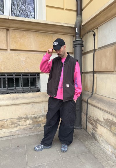 Hombre con gorra negra, sudadera rosa, chaleco negro y pantalones negros holgados, de pie en la calle de la ciudad cerca de la esquina de un edificio de piedra beige.
