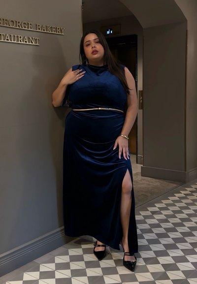 Vestido de terciopelo azul marino con una abertura lateral y un cinturón dorado, combinado con tacones negros de punta afilada. El fondo presenta un suelo de baldosas decoradas y un cartel de restaurante.