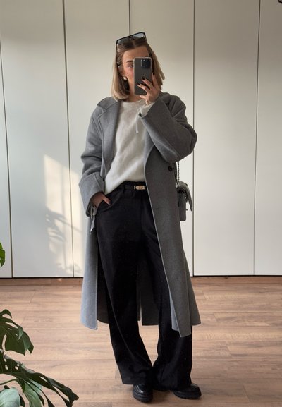 Abrigo de lana gris con cuello muesca, suéter blanco, pantalones negros y zapatos oscuros. Superficie reflectante, decoración minimalista, suelo de madera y planta verde.