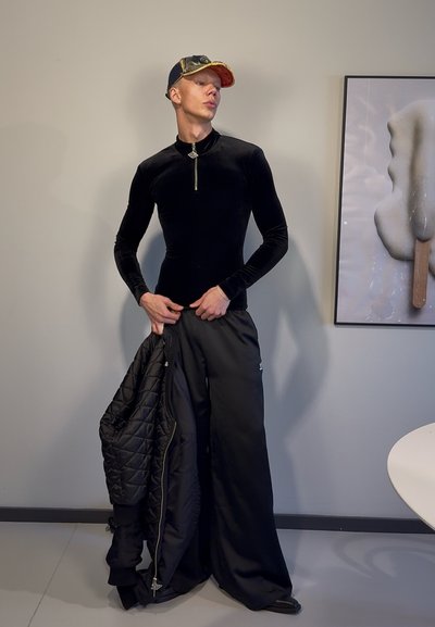 Top de terciopelo negro de manga larga con cremallera, combinado con pantalones anchos negros. Se sostiene una chaqueta negra acolchada, complementada con una gorra con visera dorada.