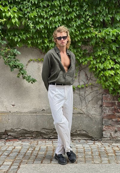 Camisa verde de botones, pantalones blancos, zapatos negros; de pie ante una pared texturizada con vegetación. Diseño minimalista y ambiente casual.