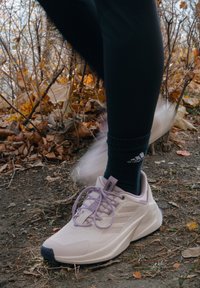 Sneaker da esterno di un leggero colore viola con tomaia texture, suola bianca e dettagli grigi. I lacci sono allacciati, indossato con calze scure e leggings neri.