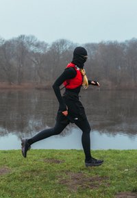 Coureur athlétique en leggings noirs et haut thermique noir, portant un gilet de course rouge, une cagoule et des chaussures noires, sur l'herbe près d'une rivière.