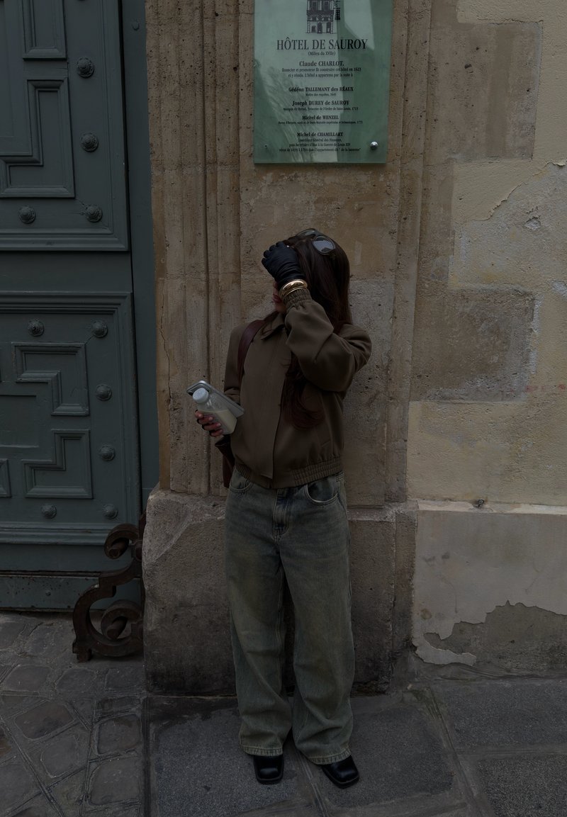 Woman in casual clothes holding a bottle, standing against a stone wall near a green door and a sign labeled Hôtel de Sauroy.