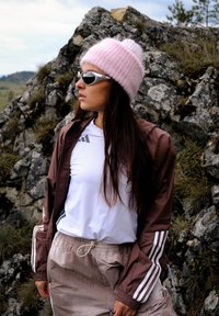 Donna con berretto di lana rosa, occhiali da sole bianchi, camicia bianca, giacca marrone e pantaloni beige che si trova all'aperto vicino a un terreno roccioso.