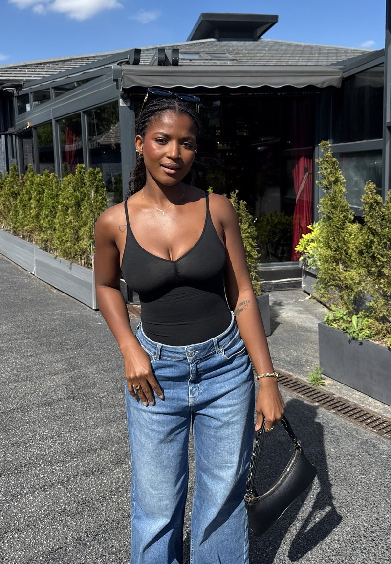 Jeune femme en débardeur noir et jean bleu tenant un sac à main noir, debout dehors près de plantes et d’un bâtiment sous un ciel bleu.