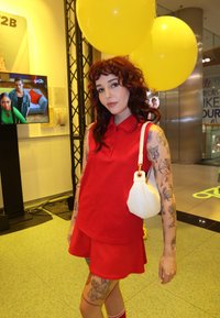 Conjunto de polo vermelho e saia, acompanhado de uma bolsa branca texturizada. Tatuagens visíveis nos braços. O fundo apresenta balões amarelos e um ecrã.
