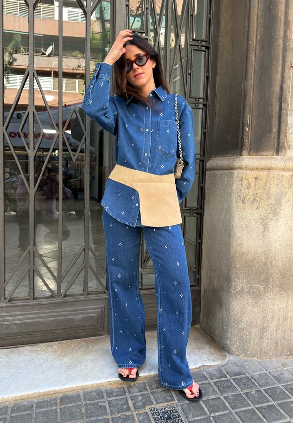 Femme portant une chemise et un pantalon en denim bleu à motif assortis avec un sac-ceinture camel, des lunettes de soleil noires et des sandales noires, debout près d'une grille en métal.
