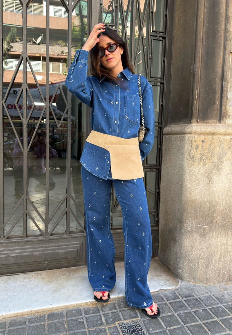 Femme portant une chemise et un pantalon en denim bleu à motif assortis avec un sac-ceinture camel, des lunettes de soleil noires et des sandales noires, debout près d'une grille en métal.