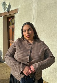 Femme aux longs cheveux noirs portant une veste marron à carreaux, tenant un sac à main noir, debout à l'extérieur près d'une porte en bois et d'un mur beige sous la lumière du soleil.