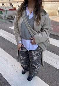 Beige Trenchcoat, grauer Pullover, weißes Hemd mit Knöpfen, graue Camouflage-Hose, schwarze Stiefel und gestapelte goldene Ringe. Gestreifter Zebrastreifen-Hintergrund.
