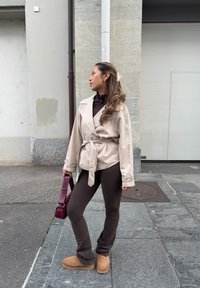 Beige omslagsfrakke med bindebælte, brune tætsiddende bukser og tanfarvede støvler. Holder en burgundy taske. Langt, bølget hår stylet med et lyst hårbånd.