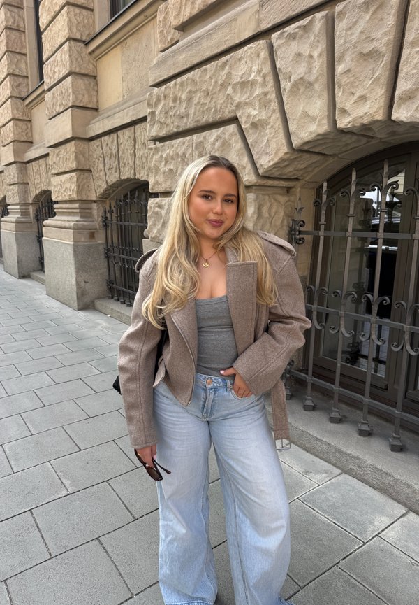 Jeune femme aux longs cheveux blonds portant une veste beige, un haut gris et un jean bleu clair se tient sur un trottoir en ville, près d'un bâtiment en pierre.