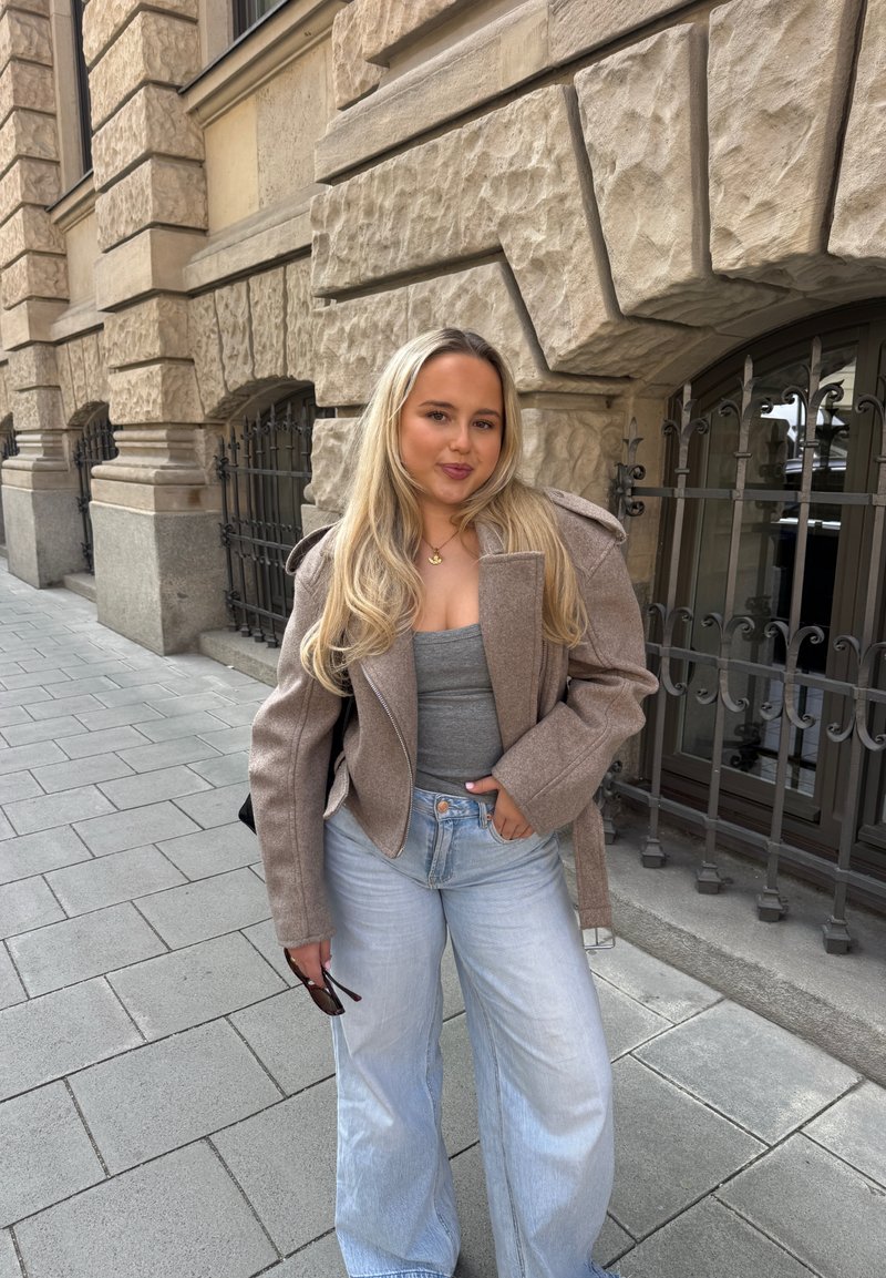 Jeune femme aux longs cheveux blonds portant une veste beige, un haut gris et un jean bleu clair se tient sur un trottoir en ville, près d'un bâtiment en pierre.