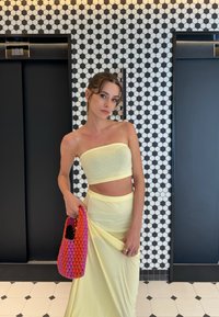 Gul tvådelad outfit med en bandeau-topp och en lång kjol. Modellen håller en vävd handväska i rosa och orange. Hexagonalt kaklat bakgrund.
