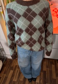 Grüner und brauner Argyle-Muster gestrickter Pullover mit geripptem Kragen. Kombiniert mit lockeren, hellblauen Denim-Jeans und schwarzen Schuhen.
