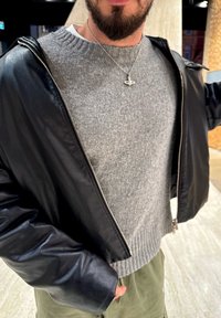 Fekete bőr rövidített dzseki világosszürke, bordázott szegélyű kötött pulóver felett, olívazöld nadrággal párosítva. Látható egy ezüst nyaklánc.