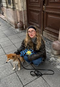 Pruun nahktagi, hall kaelarätik, sinised teksad; istub kõnniteel koos Shiba Inu koeraga; hoiab kootud sinise ja musta mustriga kotti kollase detailiga.