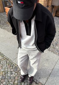 Czarna kurtka zapinana na zamek błyskawiczny z bocznymi kieszeniami, biały top z zamkiem do ćwierć wysokości, jasnoszare spodnie o szerokich nogawkach oraz ciemne buty na tle bruku.