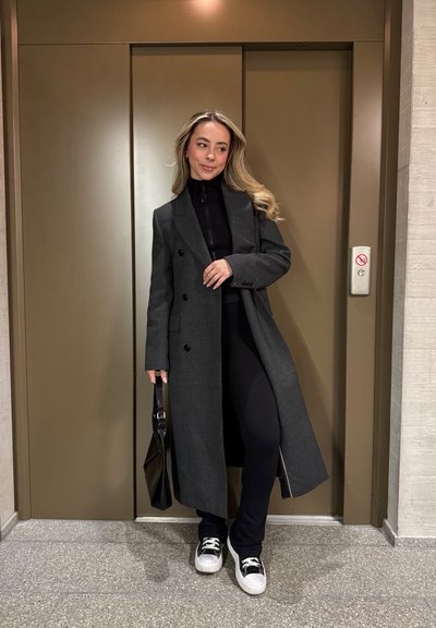 Largo abrigo gris con botones de doble pecho, combinado con una parte superior negra con cierre y pantalones. Bolsa de mano negra y zapatillas en blanco y negro. Fondo de ascensor.