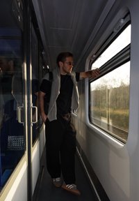 Mees seisab rongi kabiinis, kandes musta särki, tumedaid pükse ja helehalliga kampsunit õlgadel, vaadates aknast välja.