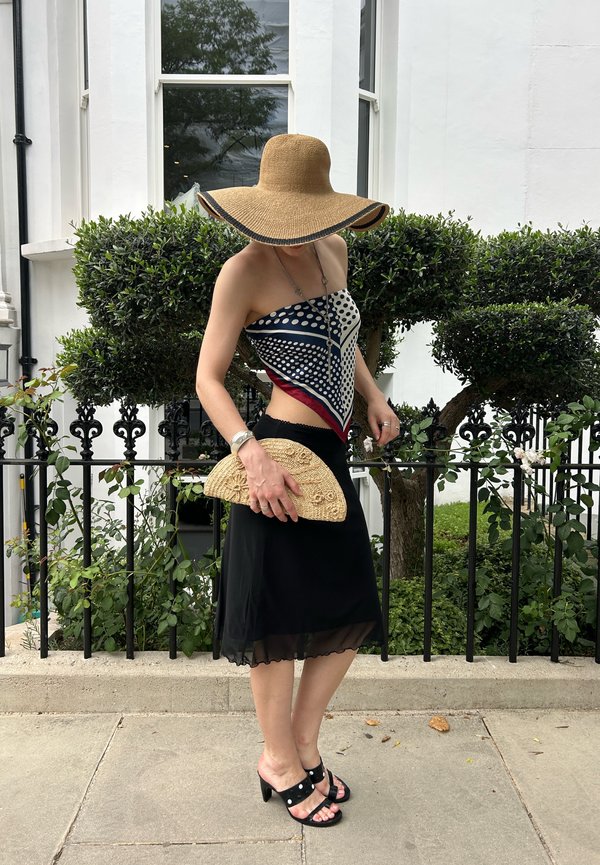 Frau mit breitkrempigem Hut, gemustertem Tuch-Oberteil, schwarzem Rock und Sandalen mit Absatz, die eine gewebte Clutch neben grünen Büschen und einem weißen Gebäude hält.
