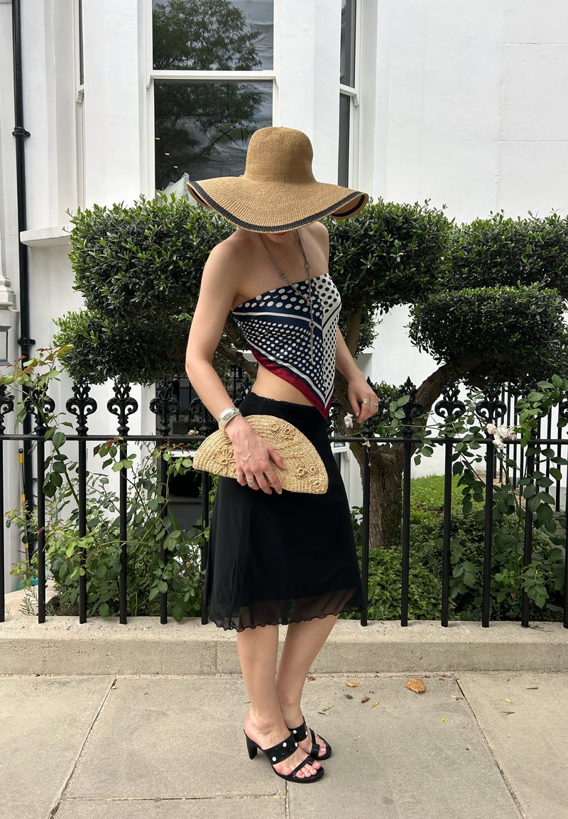 Femme portant un chapeau à large bord, un top avec foulard à motifs, une jupe noire et des sandales à talons, tenant une pochette tissée près d'arbustes verts et d'un bâtiment blanc.