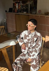 Conjunto de camisa e calças oversized em padrão castanho e branco, combinado com óculos de sol amarelos claros. Uma bolsa branca repousa sobre uma mesa de madeira.