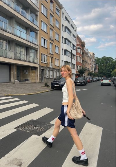 Conjunto informal que incluye una camiseta sin mangas blanca, pantalones cortos deportivos azul marino con rayas blancas, zapatos de plataforma negros y una bolsa de mimbre. Se puede ver un paso peatonal.