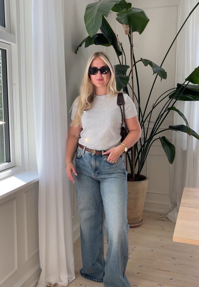Camiseta gris de manga corta, jeans de tiro alto de color azul claro, cinturón marrón, gafas de sol negras, de pie junto a una planta en maceta en una habitación luminosa.