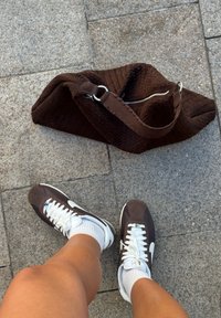 Brun vævet skuldertaske med lynlåslukning, parret med brune og hvide sneakers og hvide sokker, placeret på en stensurface.