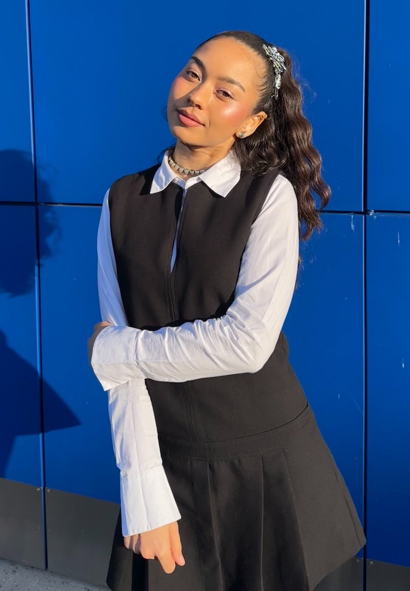 Robe noire sans manches portée sur une chemise blanche à col, avec un ourlet évasé et des plis. Le fond est d'un bleu vif. Textures légères visibles.