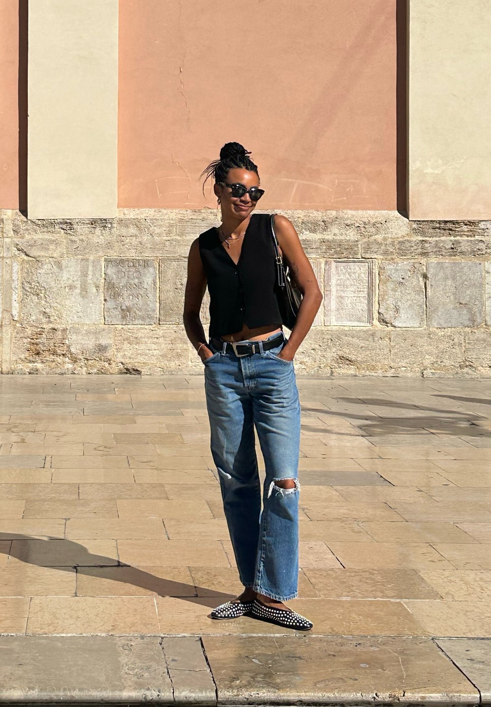 Gilet nero cropped con bottoni, jeans strappati blu chiaro, mocassini a pois bianco e nero, borsa a spalla, davanti a un muro testurizzato.