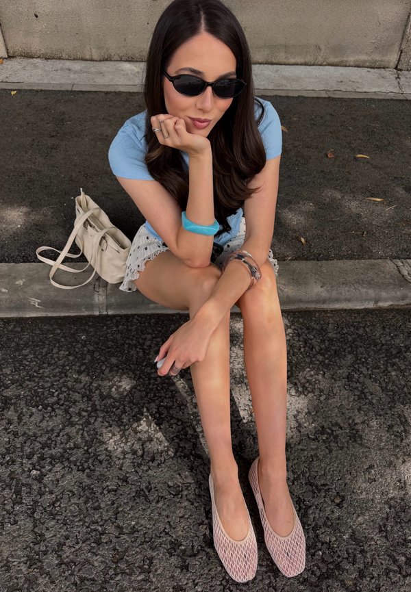 Jeune femme aux longs cheveux foncés portant des lunettes de soleil noires, un haut bleu, une jupe blanche et des chaussures roses, assise sur le bord du trottoir avec un sac beige à côté d'elle.