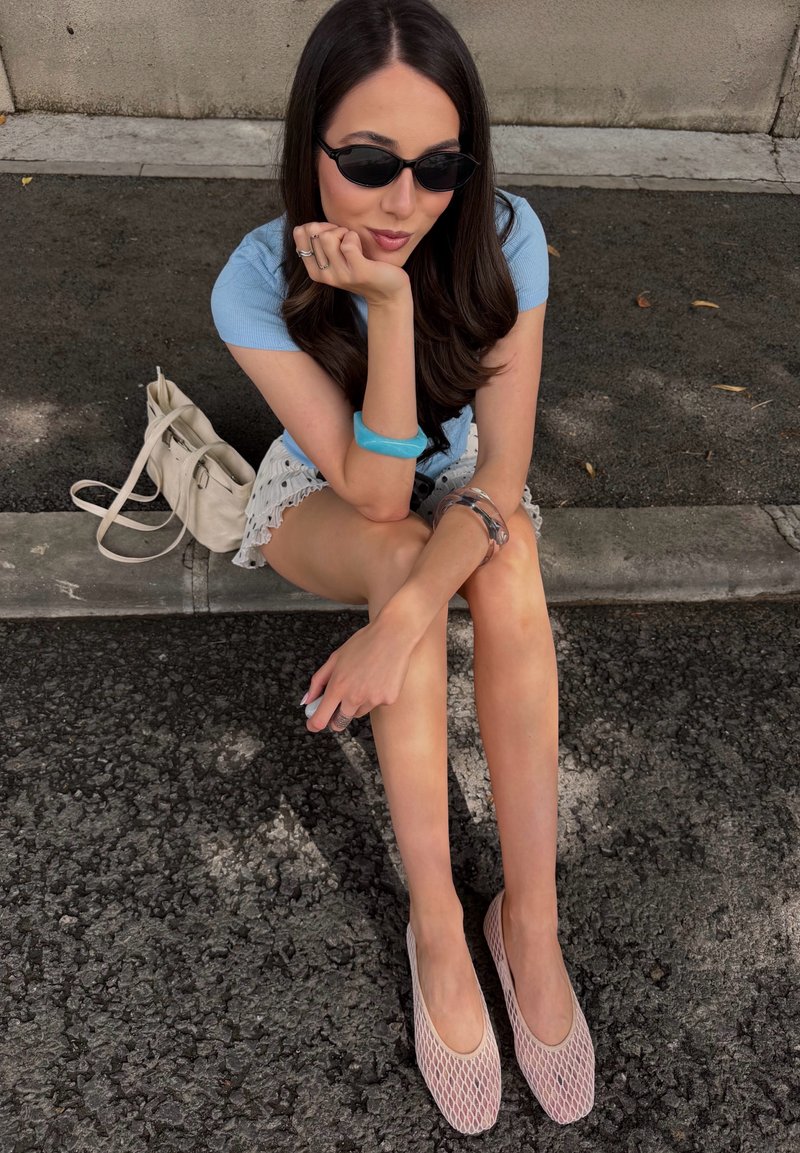 Jeune femme aux longs cheveux foncés portant des lunettes de soleil noires, un haut bleu, une jupe blanche et des chaussures roses, assise sur le trottoir avec un sac beige à côté d'elle.