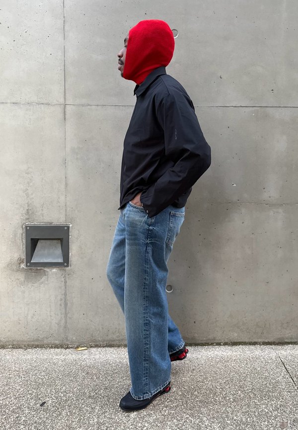 Rød strikket balaclava, sort langærmet skjorte og løse lyseblå jeans. Sorte sko med røde detaljer, baggrund med betonvæg.