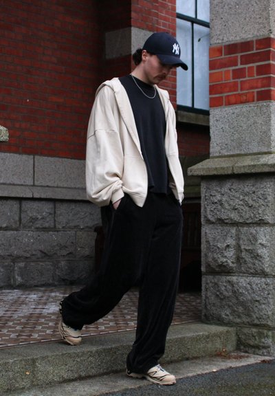 Atleta vestido con un sudadera oversize color crema, pantalones negros y zapatillas deportivas color crema. El fondo presenta una pared de ladrillo y columnas de piedra.